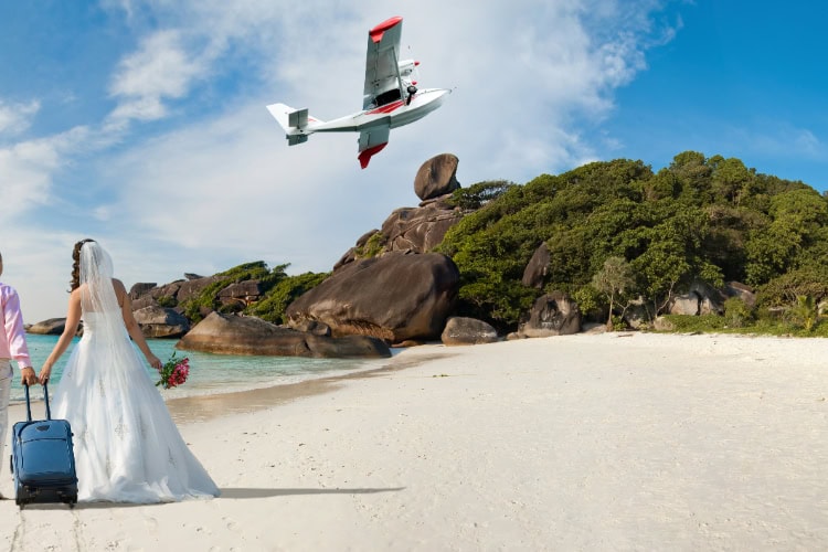 Paar steht im Anzug und Brautkleid mit einem Koffer an einem Tropischen Strand, ein Propellerflugzeug fliegt gerade davon.