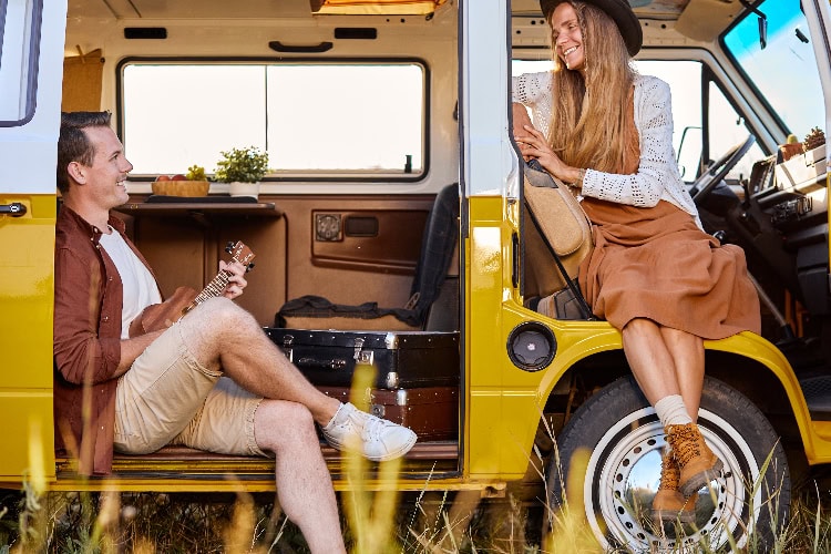 Fröhliches Paar entspannt gemeinsam am offenen Van, er spielt Ukulele, sie sitzt auf dem Beifahrersitz – entspannte Flitterwochen im Camper-Van.