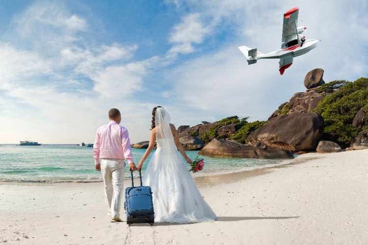 Frisch verheiratetes Paar steht mit Koffer am weißen Sandstrand, schaut einem Wasserflugzeug nach – exotischer Start in die Flitterwochen.