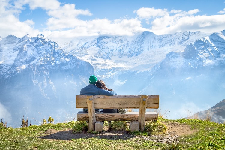 Paar sitzt auf einer Holzbank mit Blick auf schneebedeckte Berge, Arm in Arm – romantischer Moment in den Alpen.