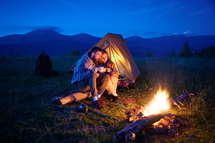 Paar sitzt abends vor einem Zelt am Lagerfeuer in den Bergen, trinkt gemeinsam aus Tassen und genießt die romantische Stimmung unter freiem Himmel.