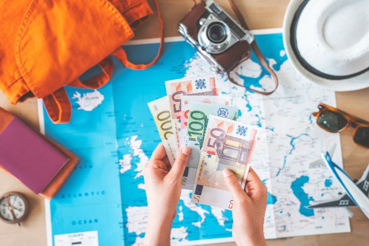 Reisende Person hält Euro-Geldscheine über einer Landkarte, daneben liegen Kamera, Hut, Sonnenbrille, Reisepass und Rucksack – Symbol für Reiseplanung und Urlaubsbudget.