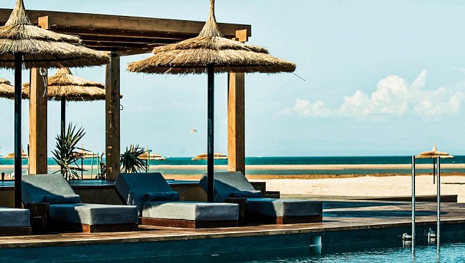 Im Vordergrund sieht man einen Teil des Pools. Direkt daran angrenzend die gepolsterten Poolliegen mit den Sonnenschirmen. Unmittelbar dahinter beginnt der Sandstrand und das Meer.