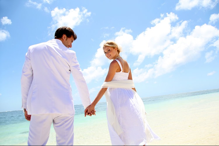 Brautpaar in weißen Outfits hält sich an der Hand und läuft am sonnigen Strand entlang; die Braut blickt lächelnd zurück