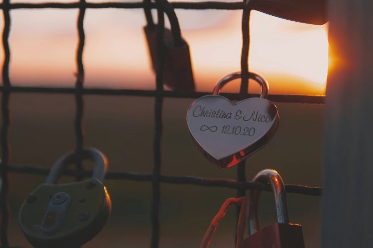 Herzförmiges Vorhängeschloss mit Gravur „Christina & Nico 12.10.20“ an einem Gitter, im Hintergrund Sonnenuntergang.