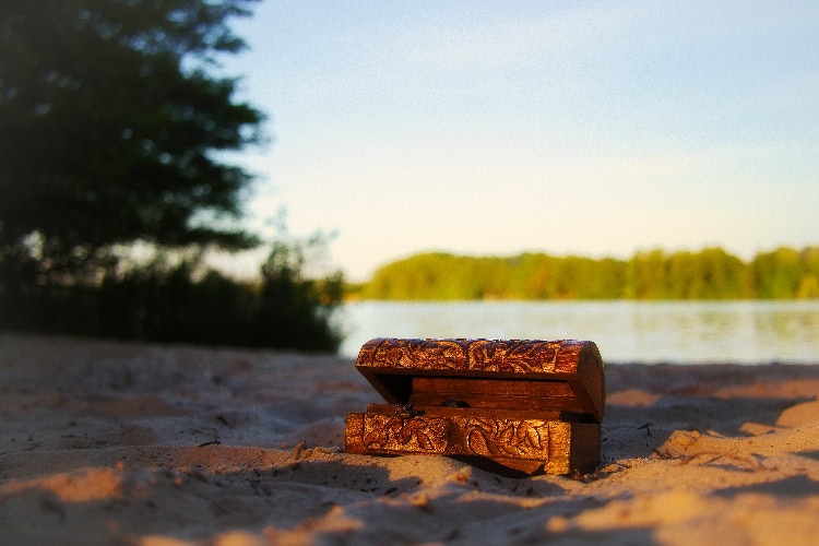 Kleine verzierte Holztruhe im Sandstrand, im Hintergrund Wasser und Bäume bei Sonnenlicht.