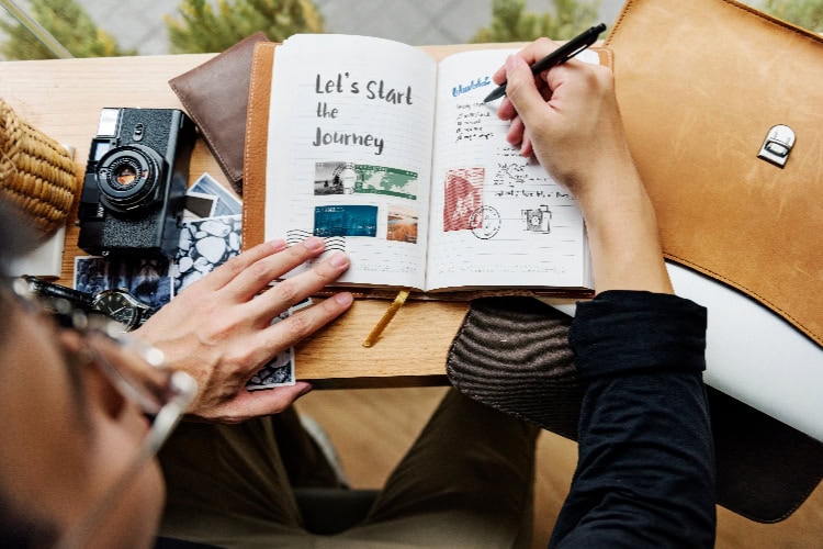 Person schreibt in ein aufgeschlagenes Reisejournal mit Fotos und Notizen, daneben Kamera und Ledertasche.