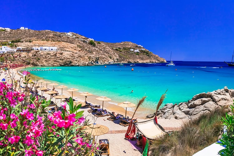 Strandaufnahme mit türkisfarbenem Wasser, Sonnenliegen und pinken Bougainvillea-Blüten an einem der beliebten Strände von Mykonos.