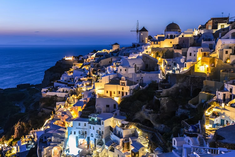 Stimmungsvolle Abendansicht von Oia auf Santorini mit beleuchteten Häusern, Windmühle und Meerblick bei Sonnenuntergang.