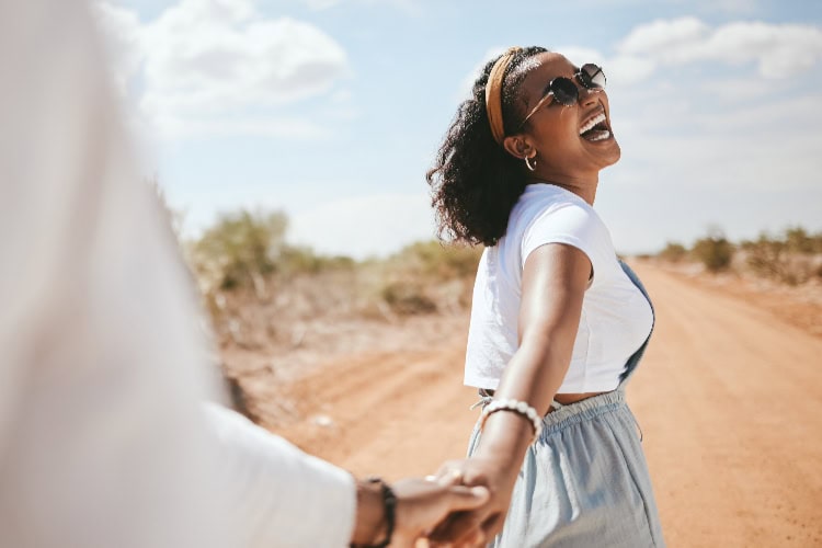 Lachende Frau auf einer staubigen Piste, die die Hand ihres Partners hält und in die Sonne blickt.