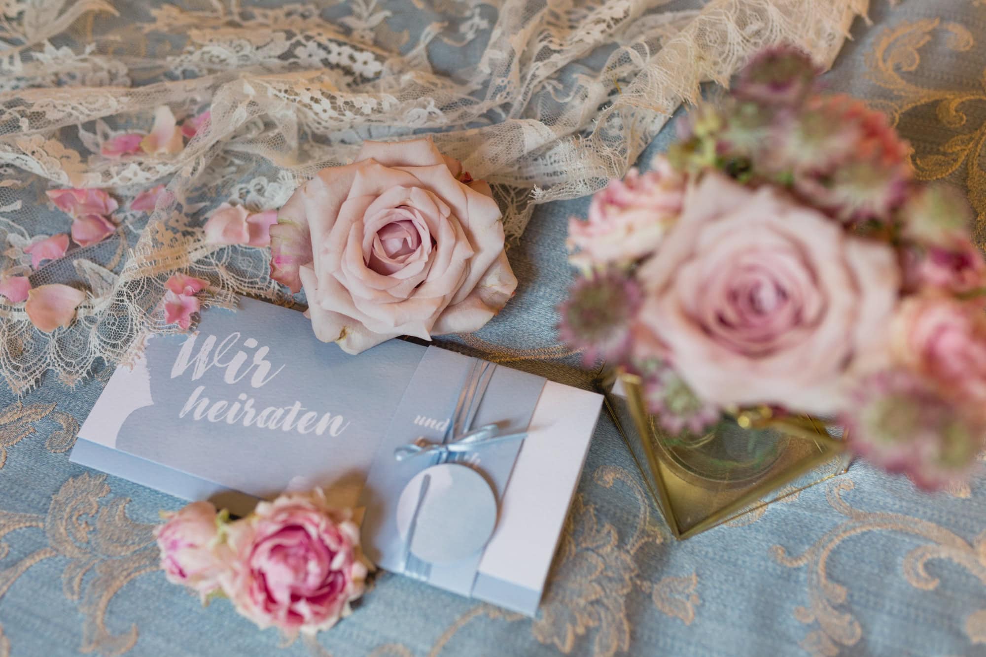 Rosa Rosen auf Spitzenstoff und blauem Hintergrund und einer Karte mit Hochzeitseinladung