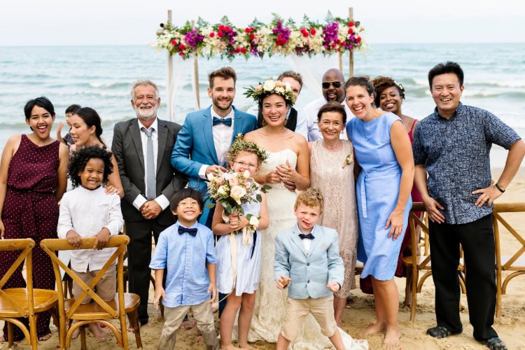 Gruppe fröhlicher Hochzeitsgäste mit Braut und Bräutigam bei einer Strandhochzeit, alle lächeln in die Kamera, Blumenschmuck und Meer im Hintergrund.