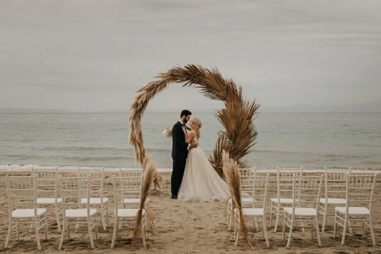 Paar steht unter Hochzeitsbogen am Meer, davor die Gästestühle