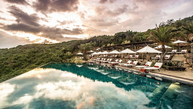 Infinity-Pool mit Sonnenliegen, Spiegelung des Abendhimmels und grünen Hügeln im Hintergrund.